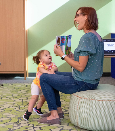 A smiling adult holds up an open book while a toddler leans on the adult's leg, looking and smiling at the book