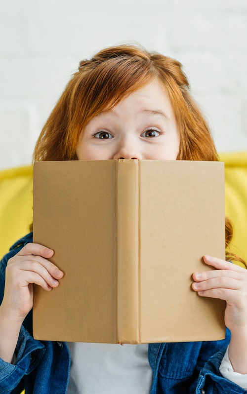 A young person peeks over an open book they are holding. Their eyes look joyful and their hair is red.