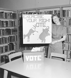 Black and white photo of a woman with voting display at the Temporary Library Headquarters, Merriam. 1956.