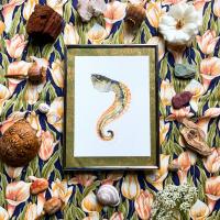 Framed watercolor painting of snake-like sea creature, set on top of floral fabric and decorated with shells and rocks.