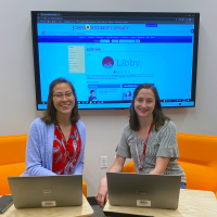 Photograph of two Librarians, Hope and Samantha, sitting on orange chairs with laptops before them and a monitor displaying a webpage behind them.