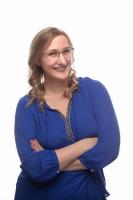 A person in a blue shirt with their arms crossed smiles at the camera. They have collarbone length blond hair.