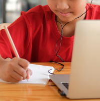 A young person in a red shirt sits at a table and writes on paper with a pencil. A laptop is open in front of them.