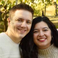 Two people, one with short brown hair and one with long brown hair, lean their heads together and smile into the camerar