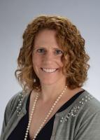 Ashley Huber woman smiling with curly shoulder length hair wearing a vneck blouse with grey sweater and long pearls