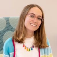 Natalie Wolf Young smiling woman with glasses, straight blondish hair, wearing a white t-shirt, sweater, and colorful necklace
