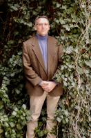 Boyd Bauman man in tan pants, brown jacket, and purple shirt standing in greenery