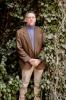 man in tan pants, brown jacket, and purple shirt standing in greenery