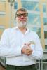 Simon Friedman Man in white button down shirt with clasped hands, smiling, black framed glasses and a white beard